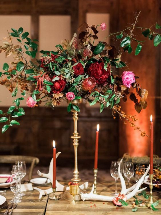 a luxurious wedding centerpiece of pink, red and burgundy blooms, greenery and dried leaves on a refined gold stand