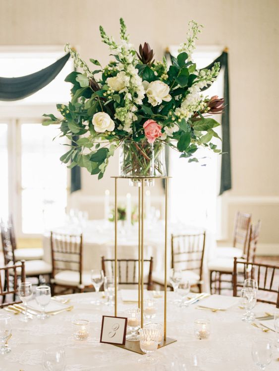 a tall and chic wedding centerpiece of white and pink blooms plus greenery on a tall gold stand