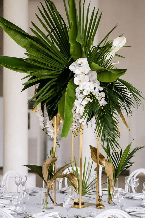 a glam tropical centerpiece of large tropical leaves and white orchids on tall gilded stands