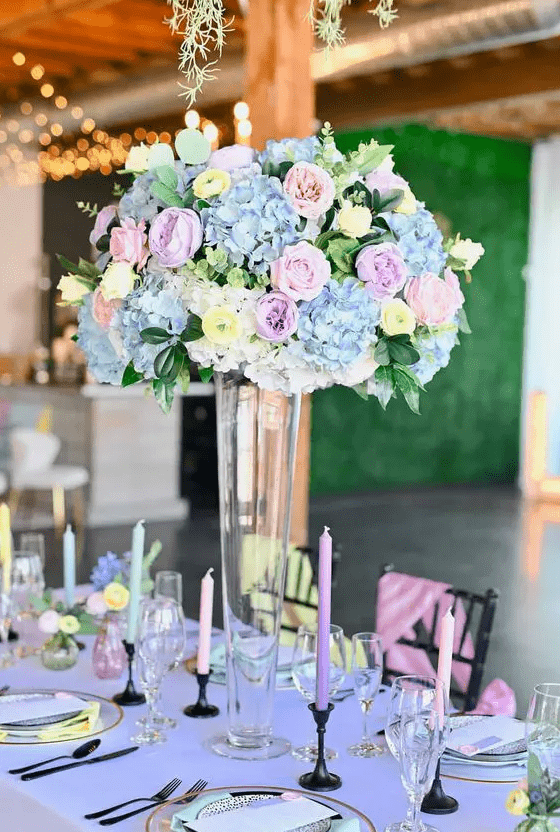 a tall pastel wedding centerpiece of blue hydrangeas, pink and lilac blooms and yellow ranunculus plus greenery
