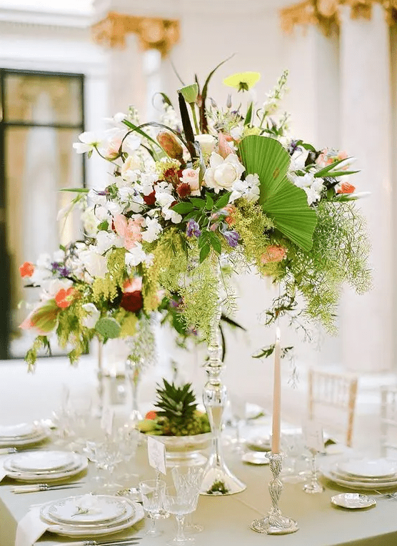a tall super lush tropical wedding centerpiece with greenery and all kinds of blooms in light colors