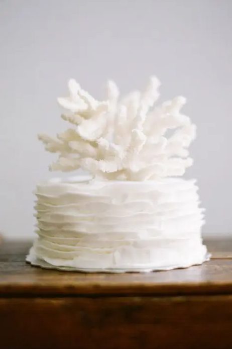 a white one-tier ruffled wedding cake with corals on top is a stylish idea for a beach wedding