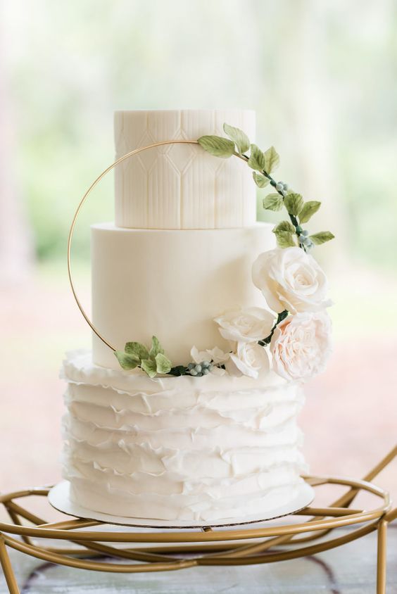a white patterned, sleek and ruffle wedding cake with a hoop and white and blush blooms is cool
