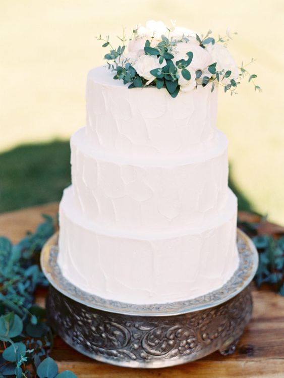 a natural and textural white wedding cake with white and blush blooms and greenery on top