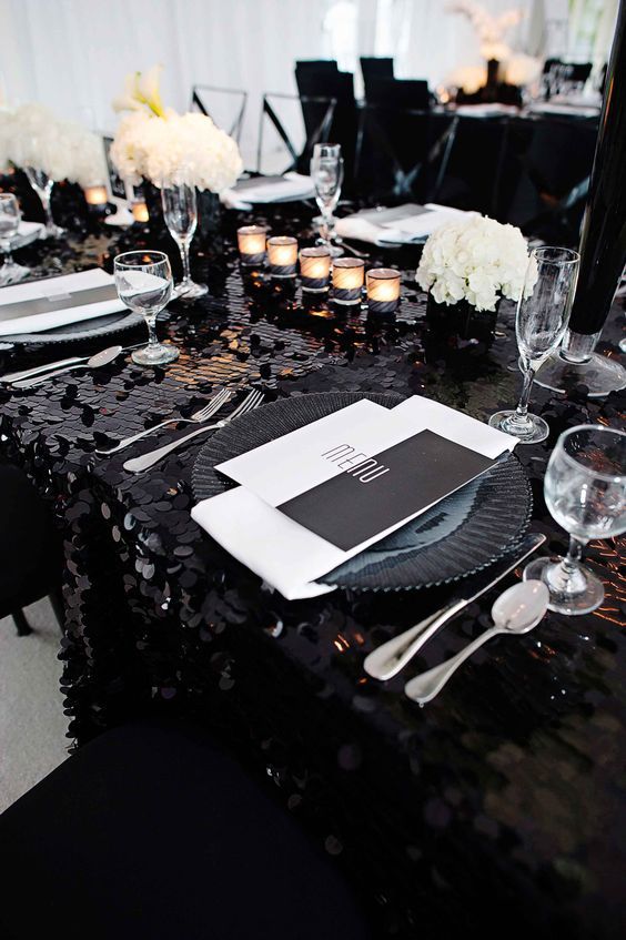 a glam wedding table setting with a black chunky sequin tablecloth, white blooms, candles, black and white stationery