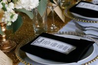 a glam wedding table setting with a gold sequin table runner, a clear charger, black napkins and white blooms and greenery