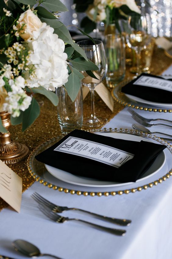 a glam wedding table setting with a gold sequin table runner, a clear charger, black napkins and white blooms and greenery