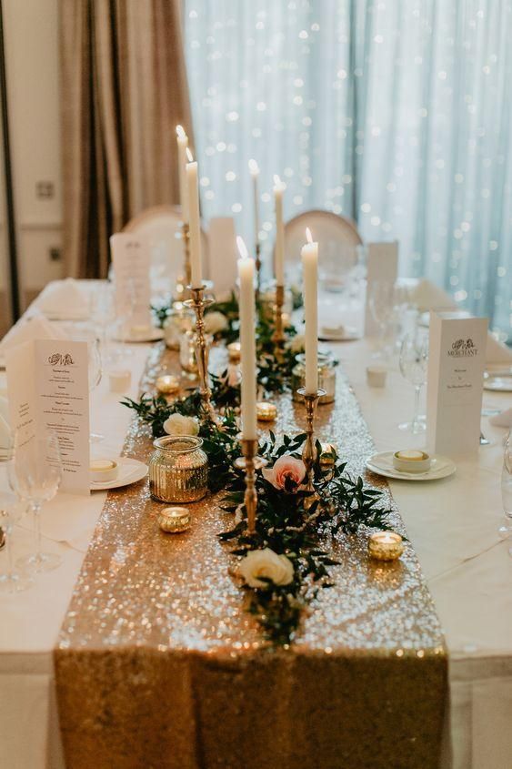 a stylish wedding tablescape with a gold sequin runner, white candles, neutral and blush blooms and greenery