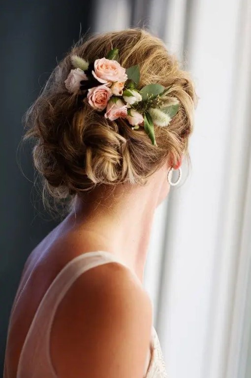 a wavy and messy low bun with a textured and dimensional top, with fresh pink roses and greenery looks classic and chic