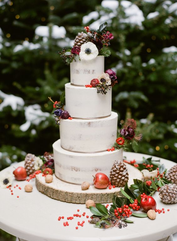a semi naked wedding cake topped with dark blooms, berries, apples and gilded pinecones