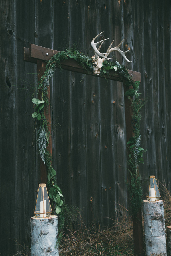 a moody Nordic wedding arch of dark stained wood, herbs and greenery, a skull with antlers and candle lanterns on each side