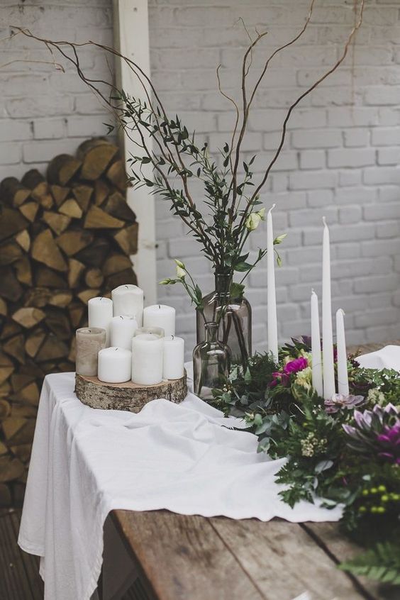 a simple centerpiece of candles on a wood slice, smoked glass vases with greenery, branches and neutral blooms