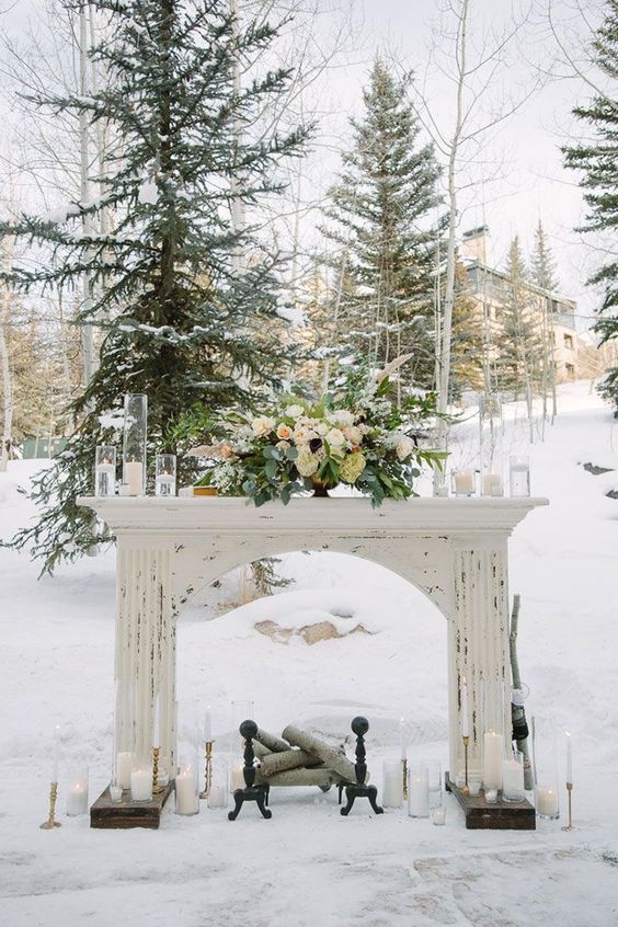 a shabby chic white mantel with candles, a lush pale centerpiece with greenery and firewood inside