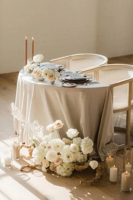 a beautiful neutral sweetheart table with a grey tablecloth, lush white blooms, tall and thin candles and grey menus