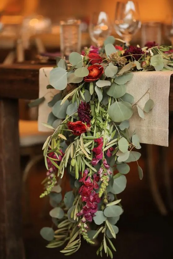 a burlap table runner, a greenery runner with red and burgundy blooms and candles for decorating a winter wedding reception