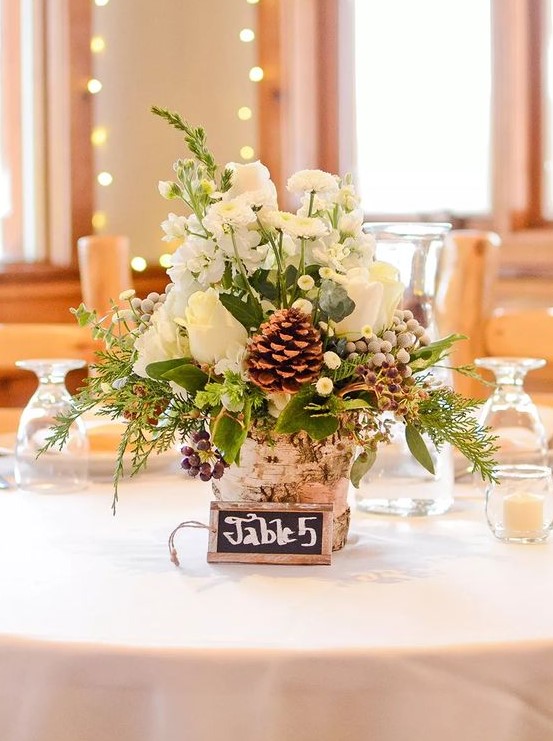 a cozy and rustic winter wedding centerpiece of a tree stump, greenery and evergreens, white blooms, berries and pinecones