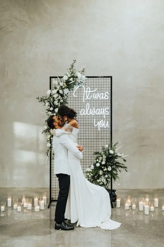 a modern winter wedding backdrop with white blooms and greenery, a neon sign and pillar candles around