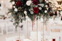 a refined tall winter wedding centerpiece with white and red roses, dahlias, greenery and evergreens, pillar candles and smaller ones