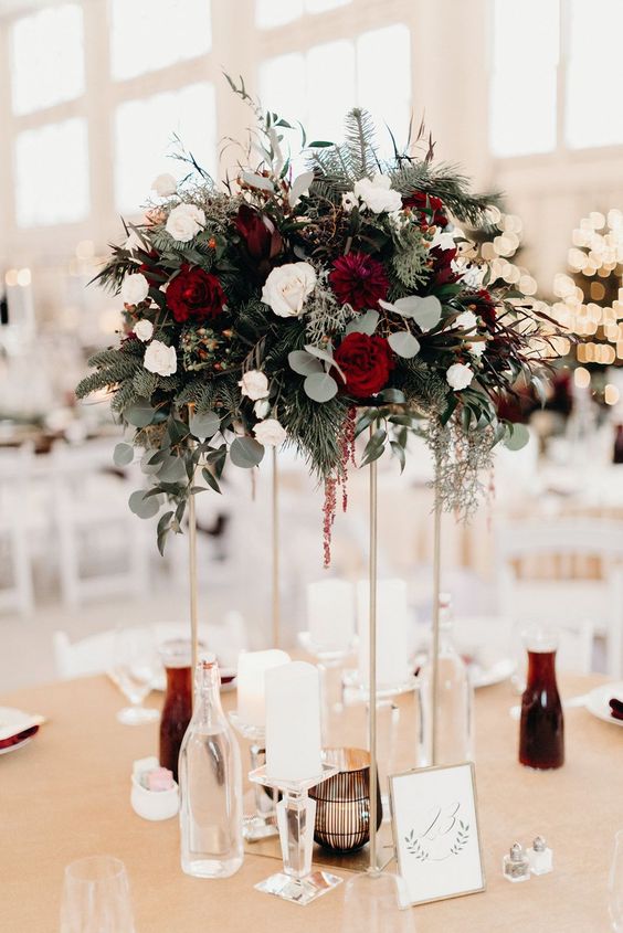 a refined tall winter wedding centerpiece with white and red roses, dahlias, greenery and evergreens, pillar candles and smaller ones