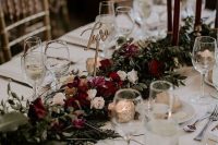 a stylish and lush winter wedding table with a refined floral and greenery runner, burgundy candles, mercury glass and an elegant table number