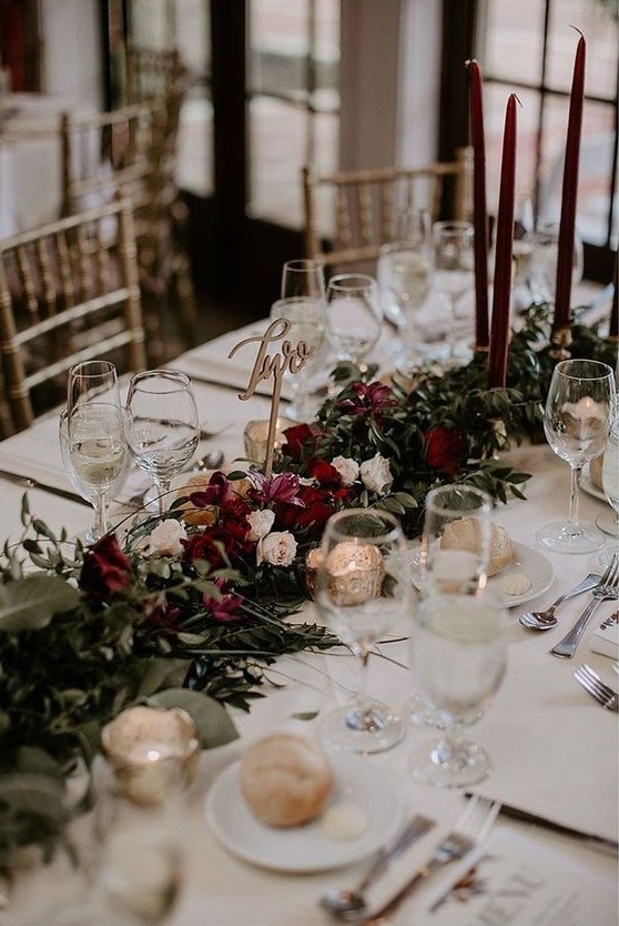 a stylish and lush winter wedding table with a refined floral and greenery runner, burgundy candles, mercury glass and an elegant table number