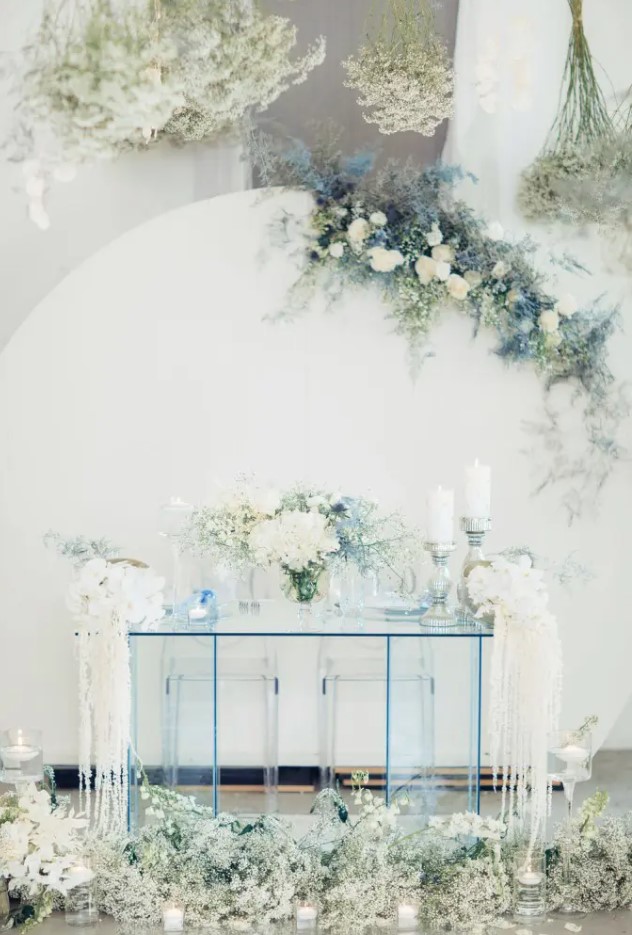 a white backdrop decorated with white and ice blue blooms and an ice blue sheer glass table and blooms for an ethereal look