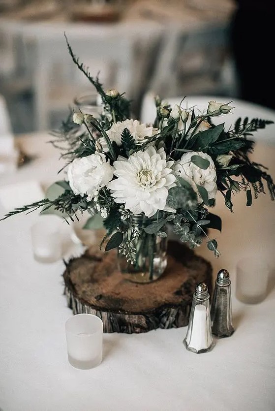 a white floral centerpiece with greenery and candles around placed on a wooden slice is a good decor idea for a winter wedding