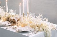 a white floral table runner of various blooms and with tall and thin candles is a refined idea for a winter wedding