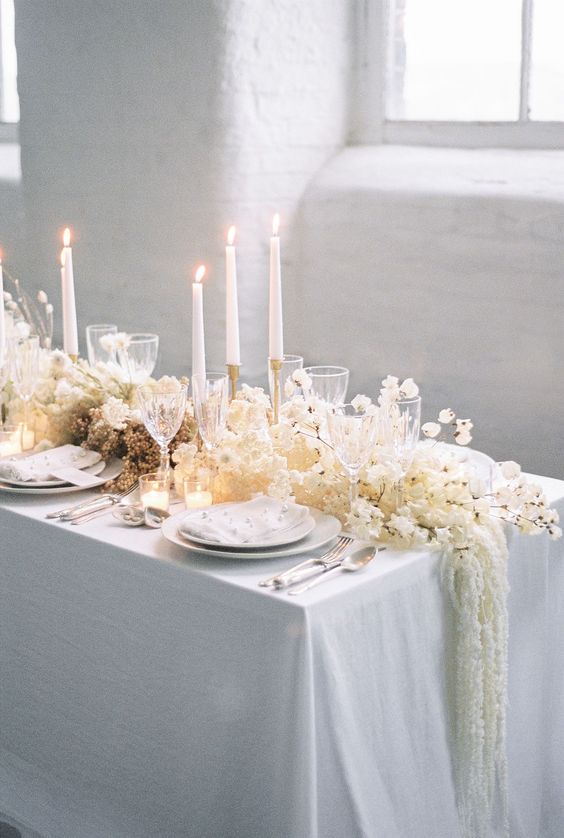 a white floral table runner of various blooms and with tall and thin candles is a refined idea for a winter wedding