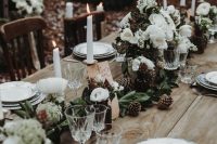 a woodland winter wedding table with an uncovered table, a greenery and white bloom runner, pinecones, white candles and printed plates