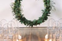 a minimalist Nordic wedding ceremony space with a giant greenery wedding wreath, sheer chairs and candles lining up the aisle