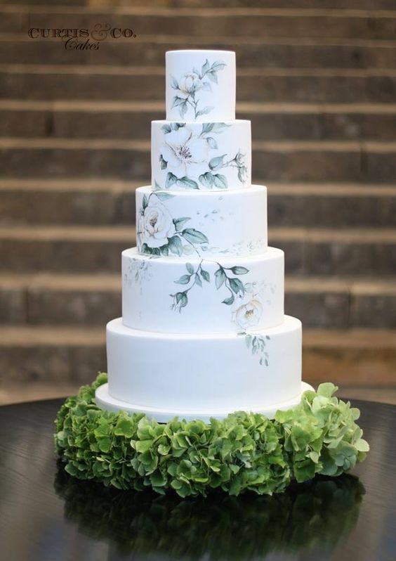 a beautiful and neutral handpainted wedding cake with several floral tiers and a white one plus a hydrangea pillow