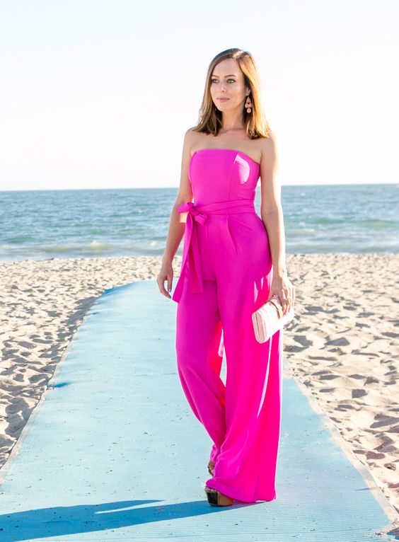 a hot pink strapless jumpsuit with a sash, a small clutch and pink earrings for a modern look