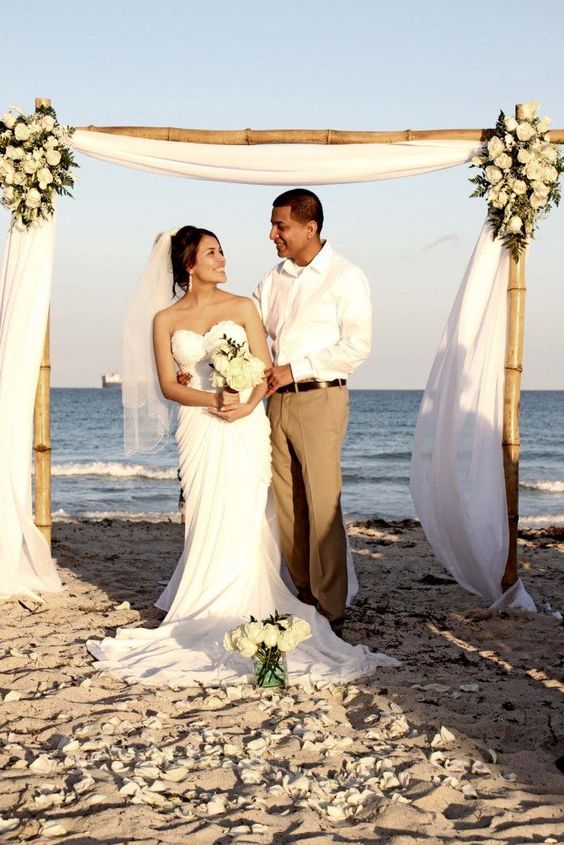 a bamboo wedding arch decorated with white flowy fabric and lush white blooms on the corners