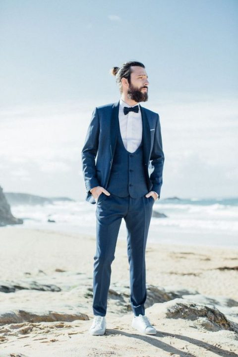 a navy three-piece suit with a white shirt, a navy bow tie and white sneakers is a chic coastal groom outfit