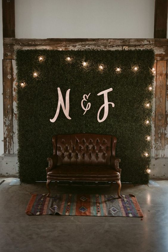 a simple wedding photo booth with a greenery wall, lights, a brown leather seat and a colorful rug
