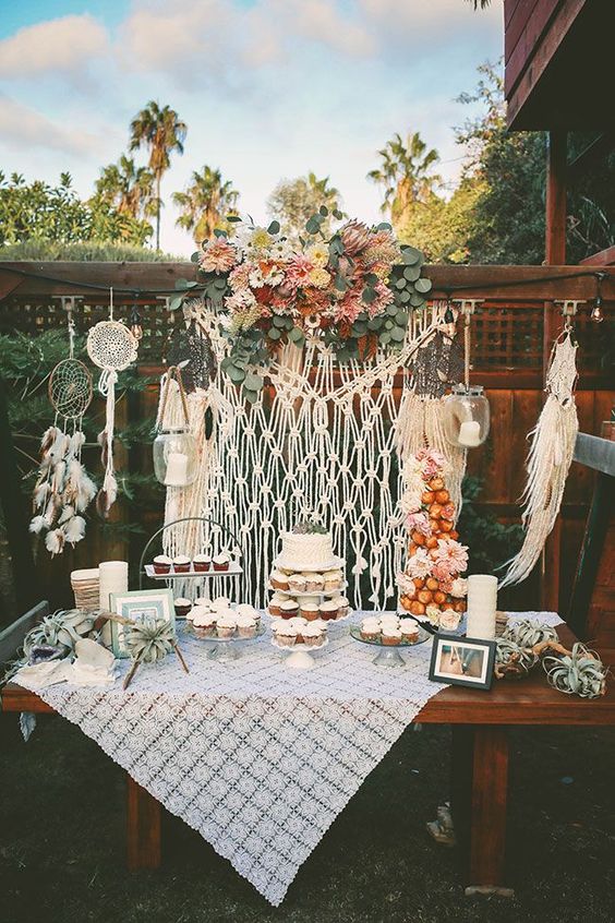 a boho backyard dessert table with a macrame backdrop and dream catchers, a crochet tablecloth and bold blooms