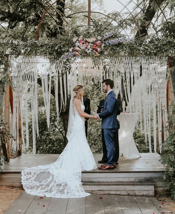 a chic macrame wedding backdrop with long fringe, lush greenery and pink blooms on top for a summer wedding