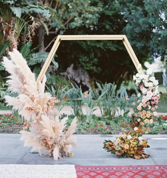 a hexagon wedding arch decorated with pampas grass and pink blooms on one side and ombre blooms on the other side, from whiet to rust