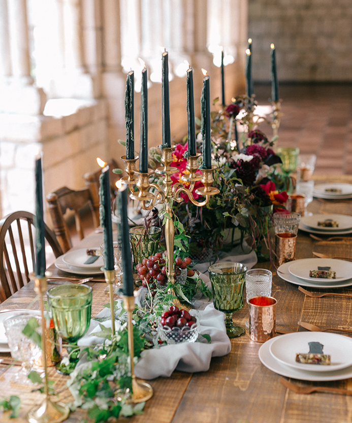 a luxurious wedding tablescape with bold lush florals, black candles in gold candle holders, greenery, fruits and berries plus copper mugs