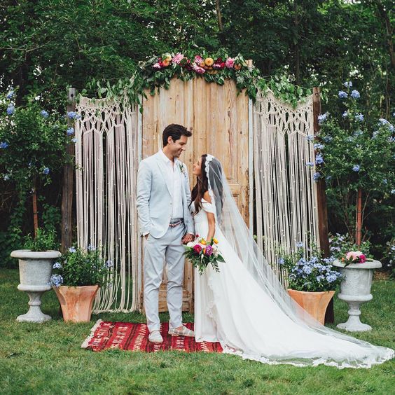a wedding backdrop made of vintage doors, macrame and some greenery and bright flowers on top