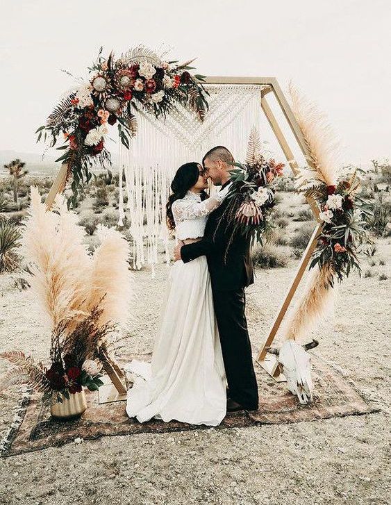 a hexagon wedding arch with a macrame hanging, pampas grass, lush white and pink blooms and foliage for a boho wedding