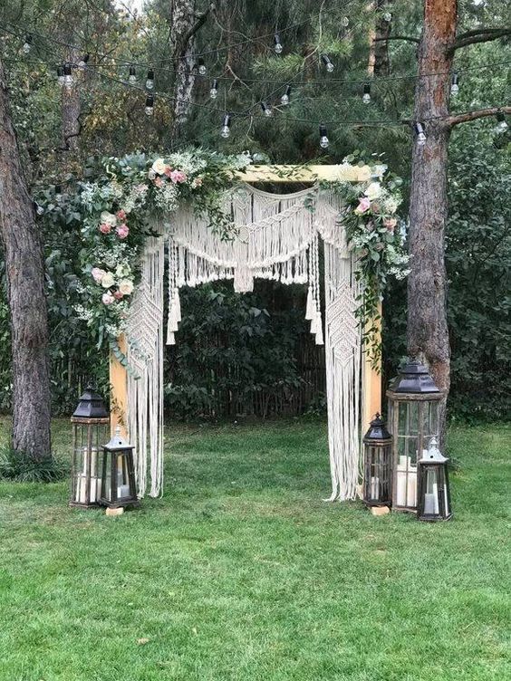 a boho macrame wedding arch with lush blooms and greenery on top plus candle lanterns around