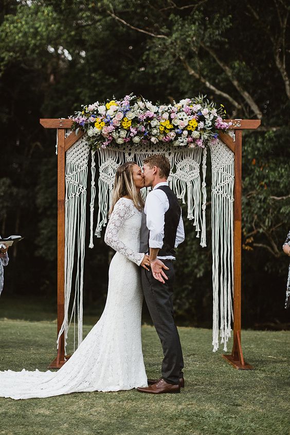 a bright boho summer wedding arch with macrame and lush blooms in pink, lilac and yellow on top