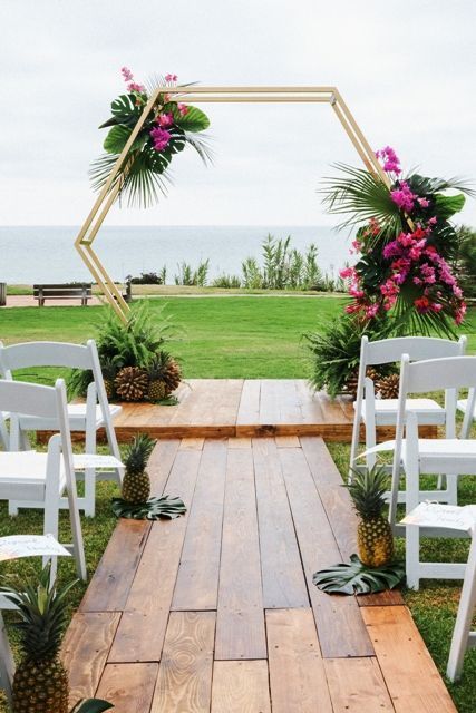 a tropical hexagon wedding arch decorated with tropical leaves, bright pink florals and tropical fruits at the base