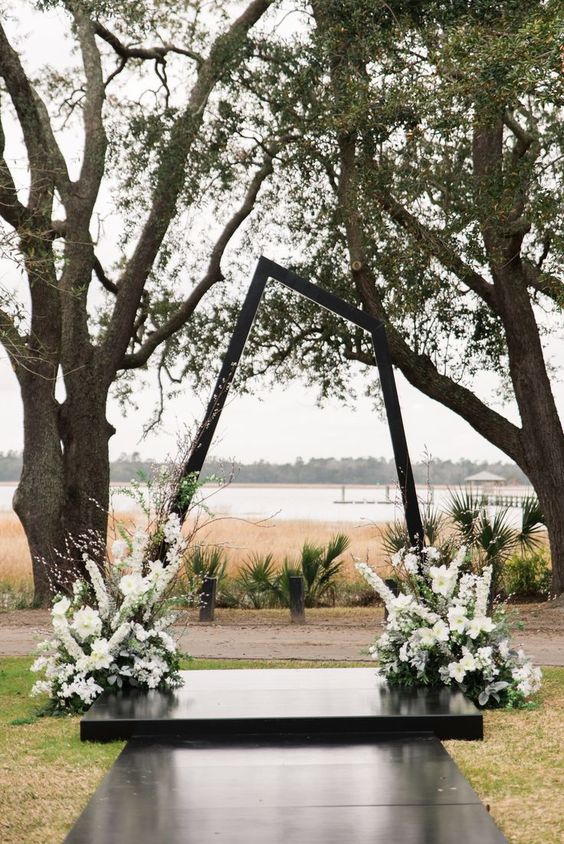 a dark stained asymmetrical geometric wedding arch with lush white blooms and greenery at its base