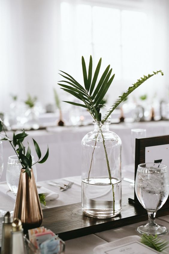 a minimalist wedding centerpiece with greenery in a clear and metallic vase on a dark stained board