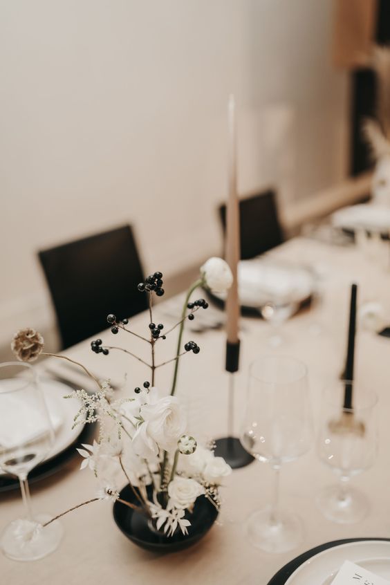 a minimalist wedding centerpiece of a black bowl, white blooms, a seed pod and berries and a tall and thin candle