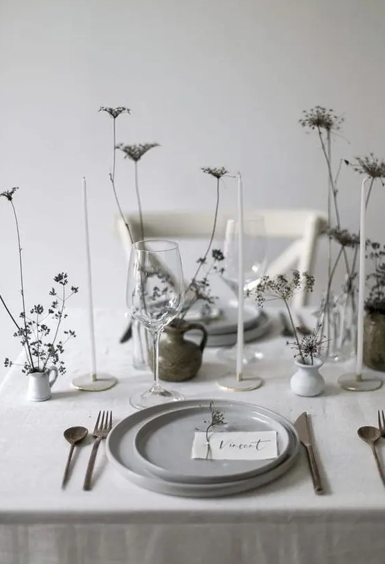 a neutral minimalist wedding tablescape with grey plates, white vases with dried blooms, simple cutlery and thin and tall candles