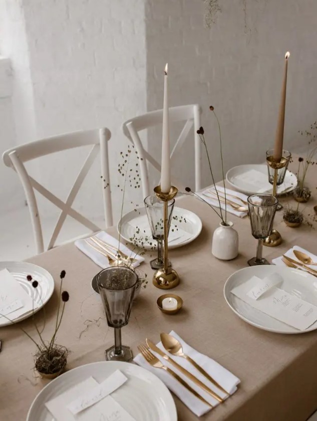 a warm-colored minimalist winter wedding tablescape with a tan tablecloth, gold touches, grey glasses, dried flowers and all the rest done in white
